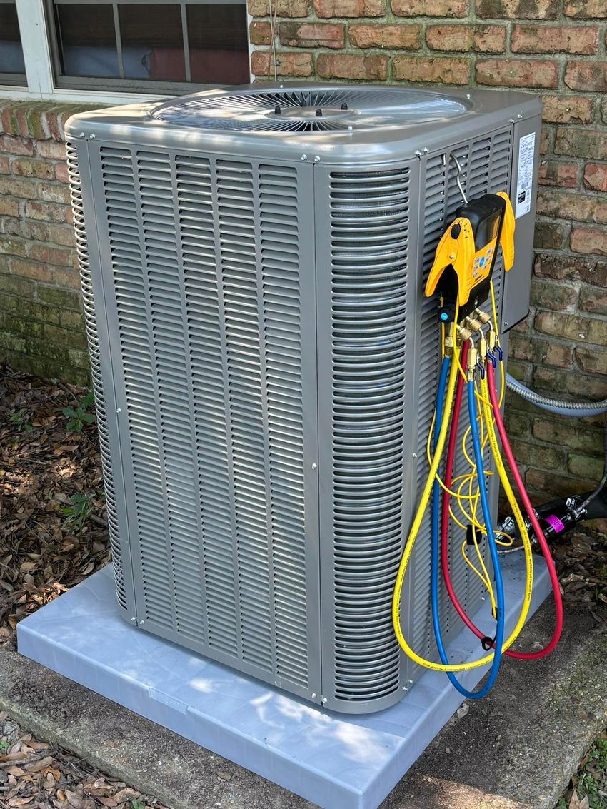 Air conditioning unit with diagnostic gauges attached, sitting on a gray pad next to a brick wall.