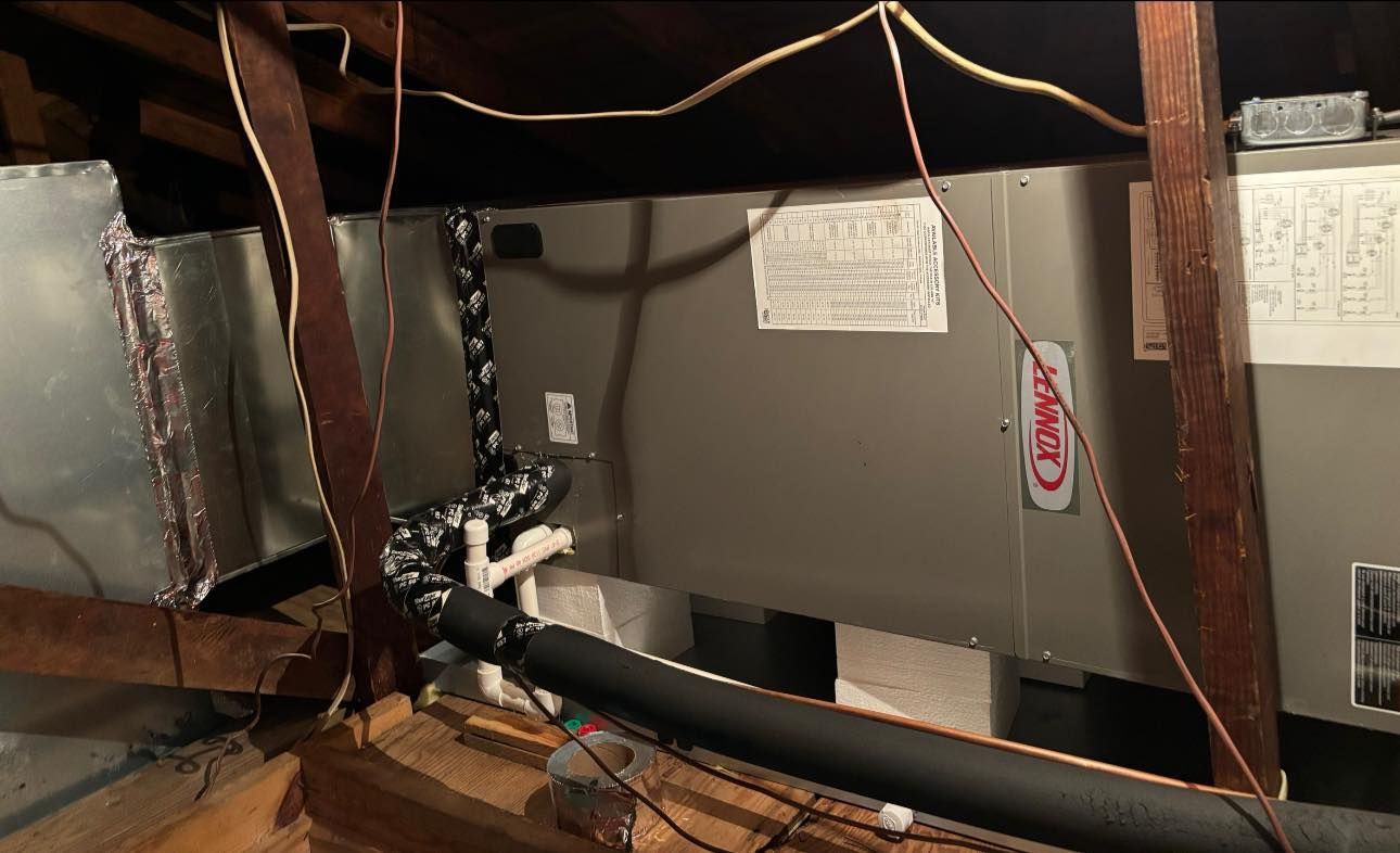 HVAC system in an attic. Metal ducts and insulated lines are visible. Brown wooden beams are overhead.