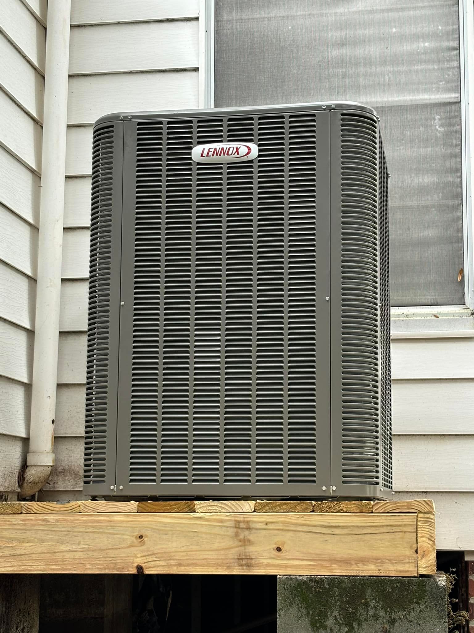 Gray Lennox air conditioner unit on wooden platform, beside white siding and window.