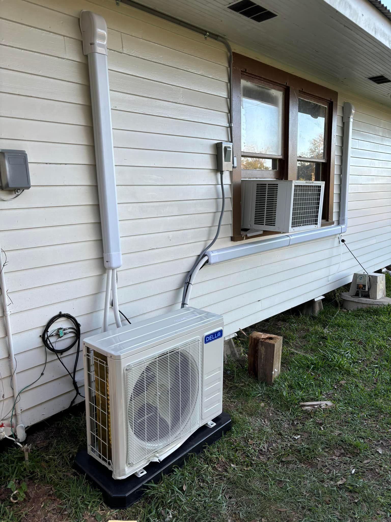 Outdoor HVAC unit with white siding, window unit, and connecting cables.