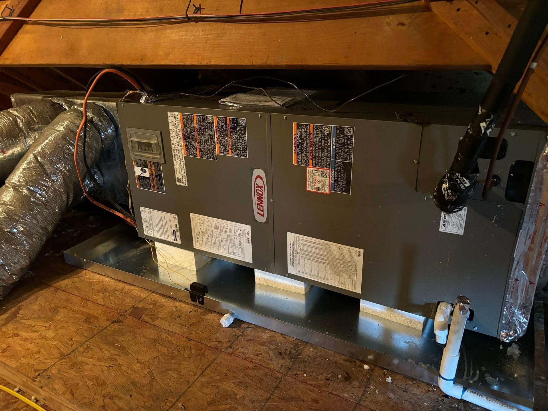 HVAC unit in an attic. Gray metal box with ductwork and pipes, mounted on a metal platform. Wooden beams overhead.