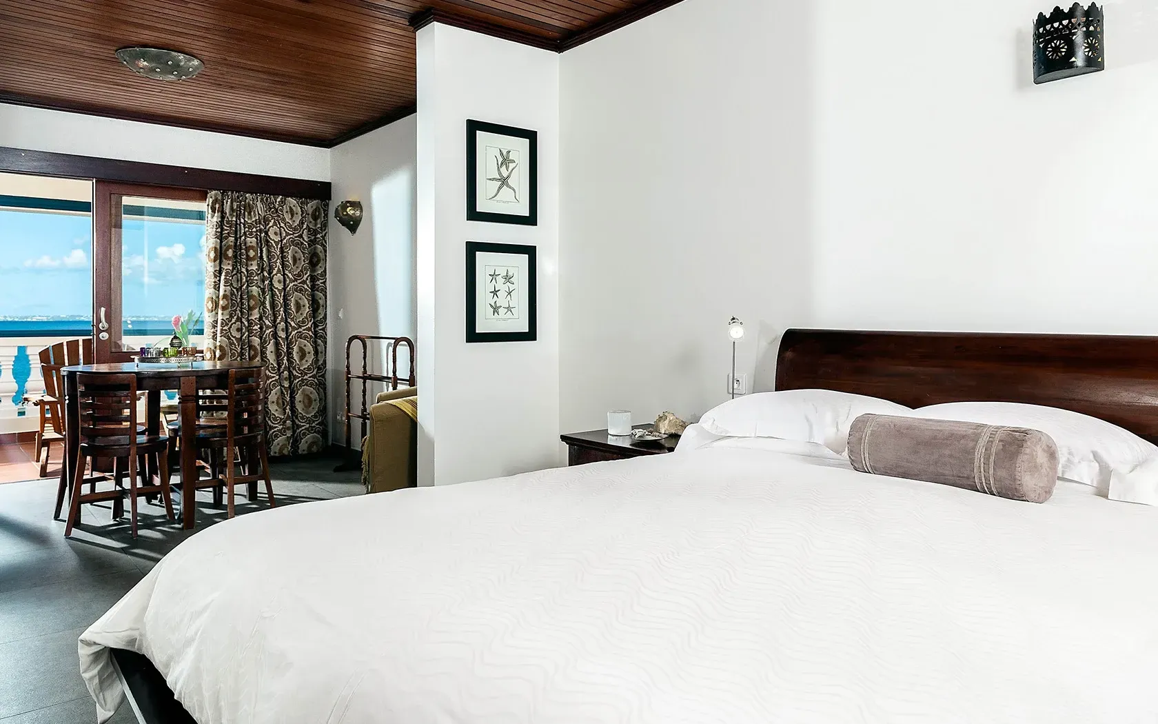 Bedroom with balcony view of ocean and dining table; bed in foreground.