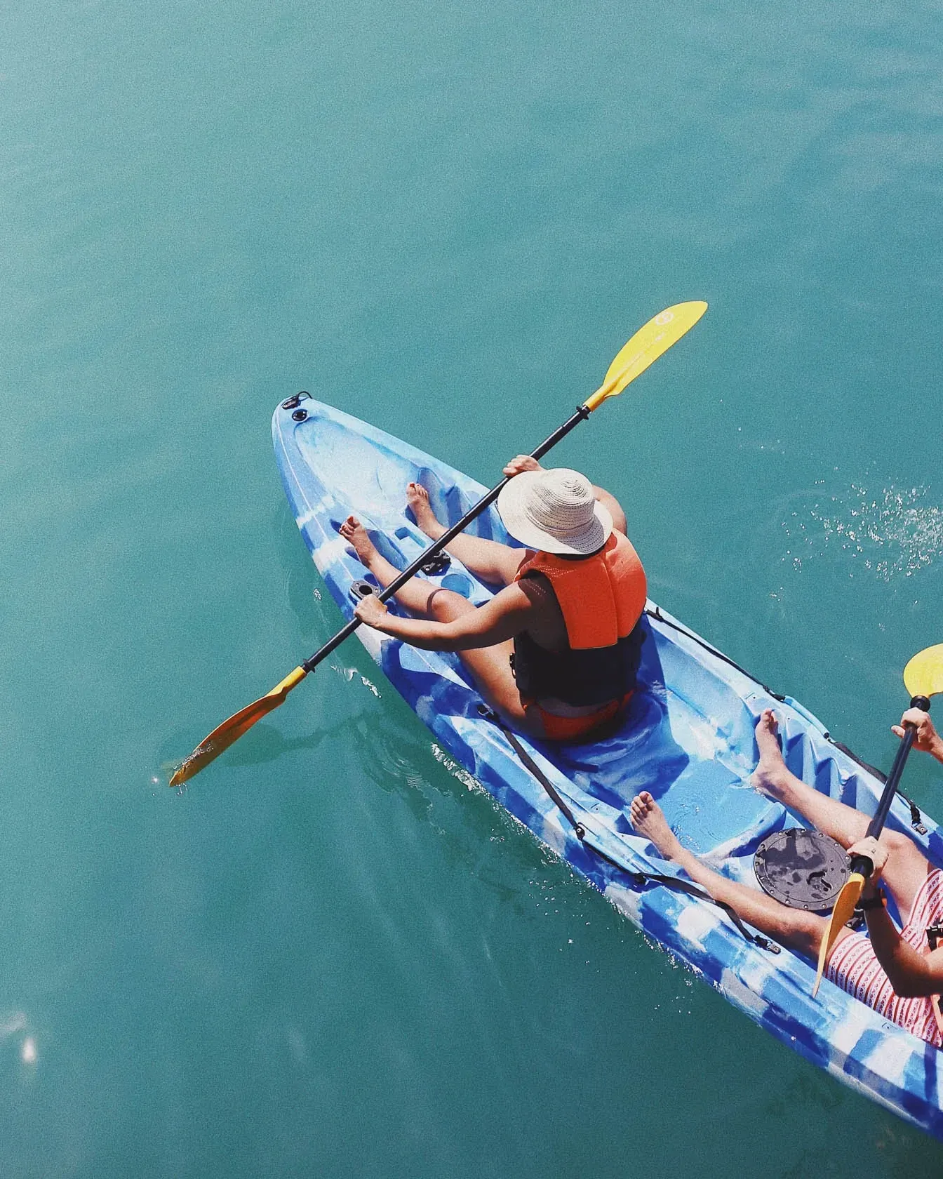 Two people kayaking on turquoise water. One wears a life vest and hat.