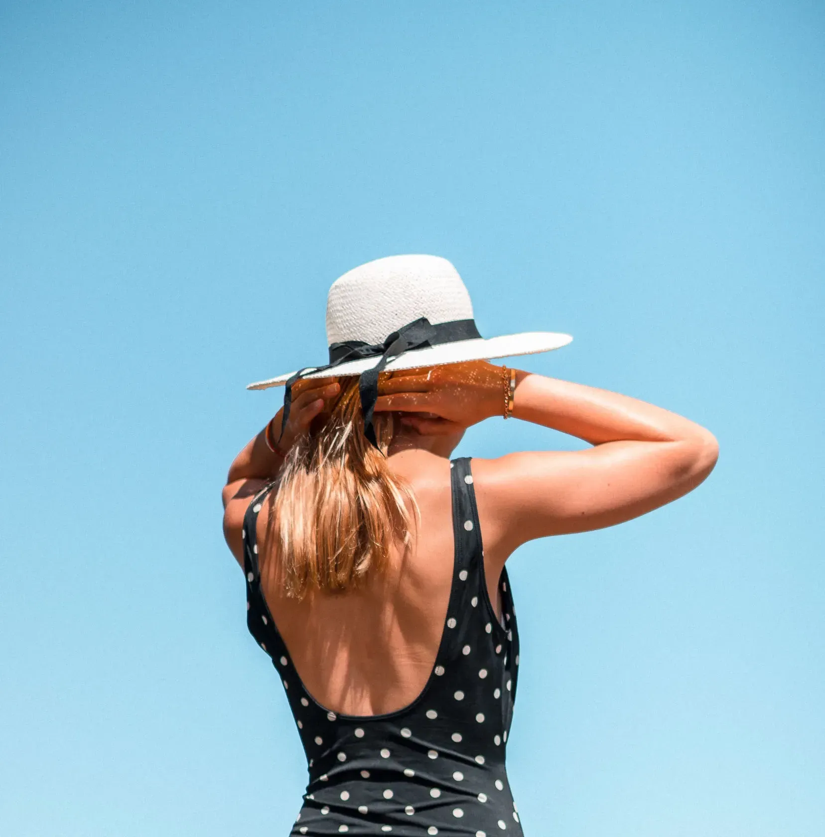 Woman in black polka dot swimsuit and sun hat against a clear blue sky.