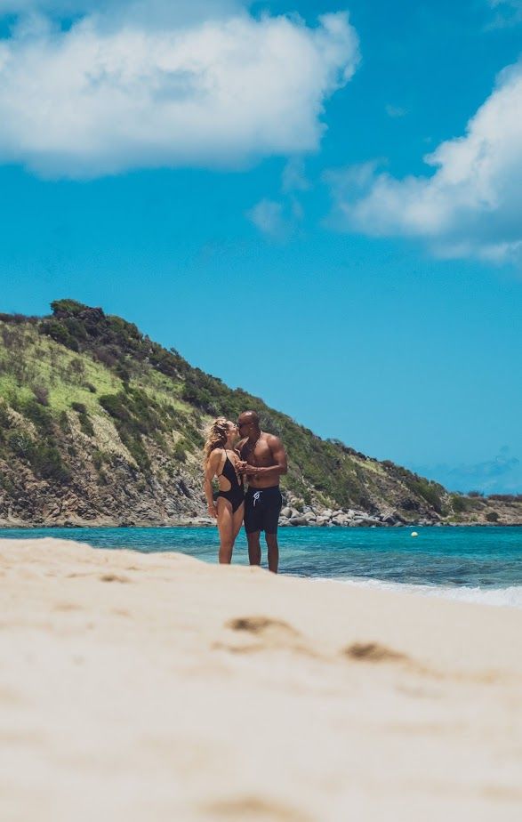 Couple standing on a beach, embracing. Turquoise water, blue sky with clouds. Hilly, green landscape in the background.