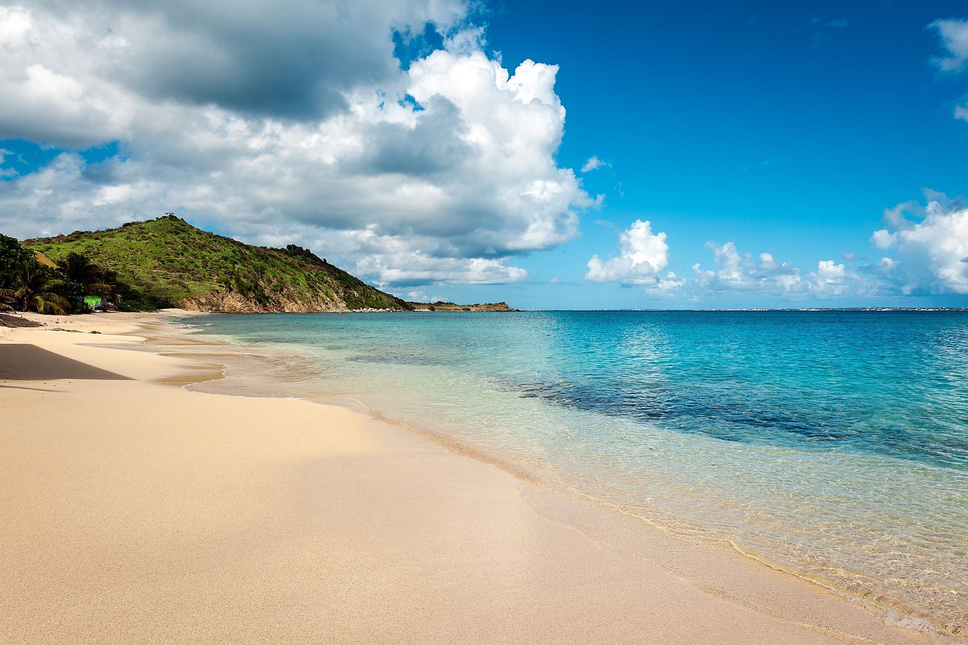 Sandy beach with clear turquoise water, bright blue sky, and green hills in the background.