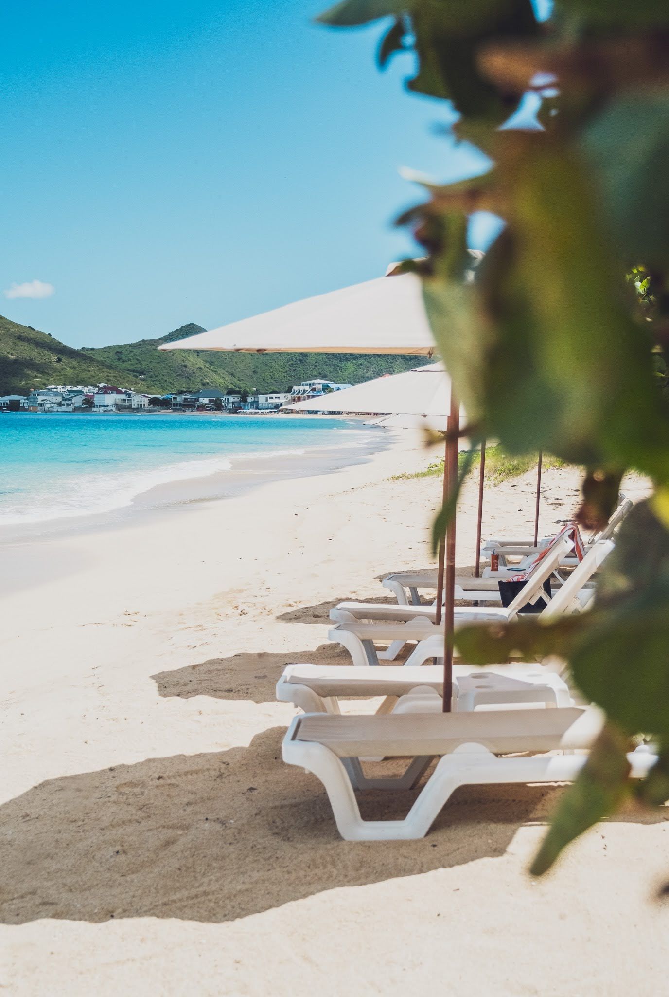 Beach scene with white sand, turquoise water, beach chairs under umbrellas, and lush green foliage.
