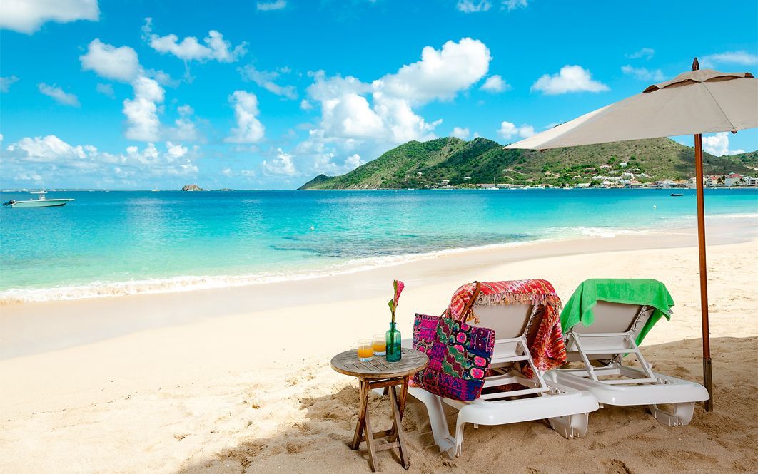 Beach scene: lounge chairs under umbrella, turquoise water, blue sky, small table with items, distant island.