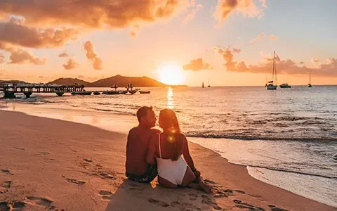 Couple on beach watching sunset; boats in water, pier in background, orange sky.