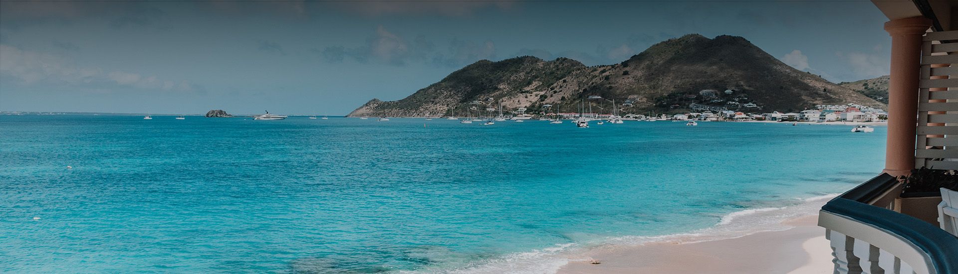 Beach with turquoise water, white sand, and mountains in the distance. Boats and balcony.