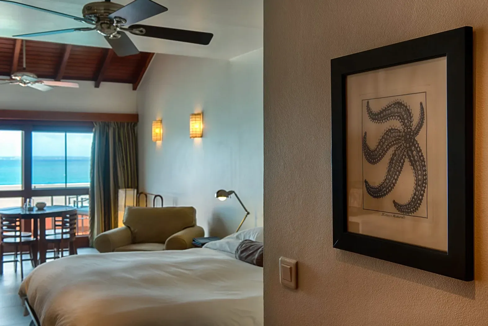 Hotel room with ocean view, framed octopus art, bed, chair, lamp, ceiling fan.