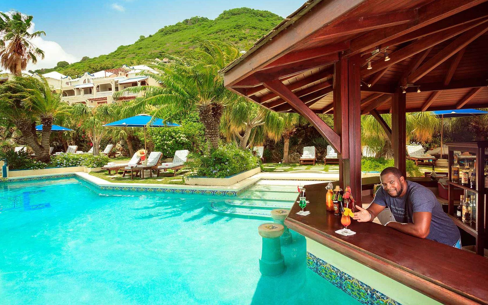 Poolside bar with bartender; resort setting with pool, lounge chairs, and tropical foliage.