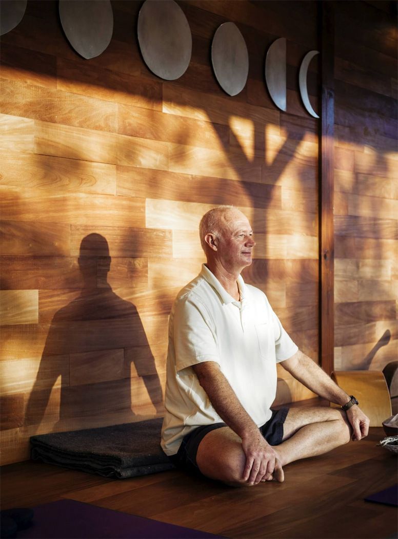 An older man sits in a meditation pose with a shadow against a wooden wall.