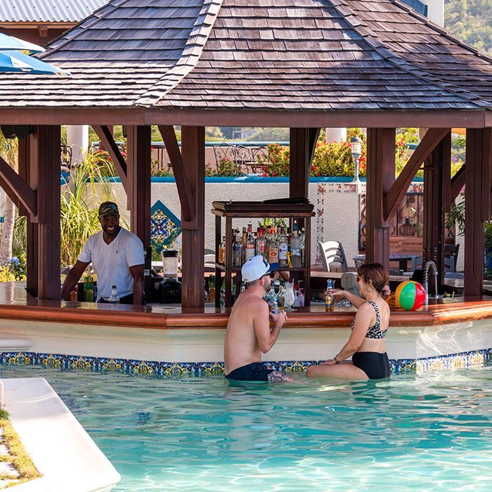 Poolside bar: patrons sit in water, bartender makes drinks, wooden gazebo, tropical setting.