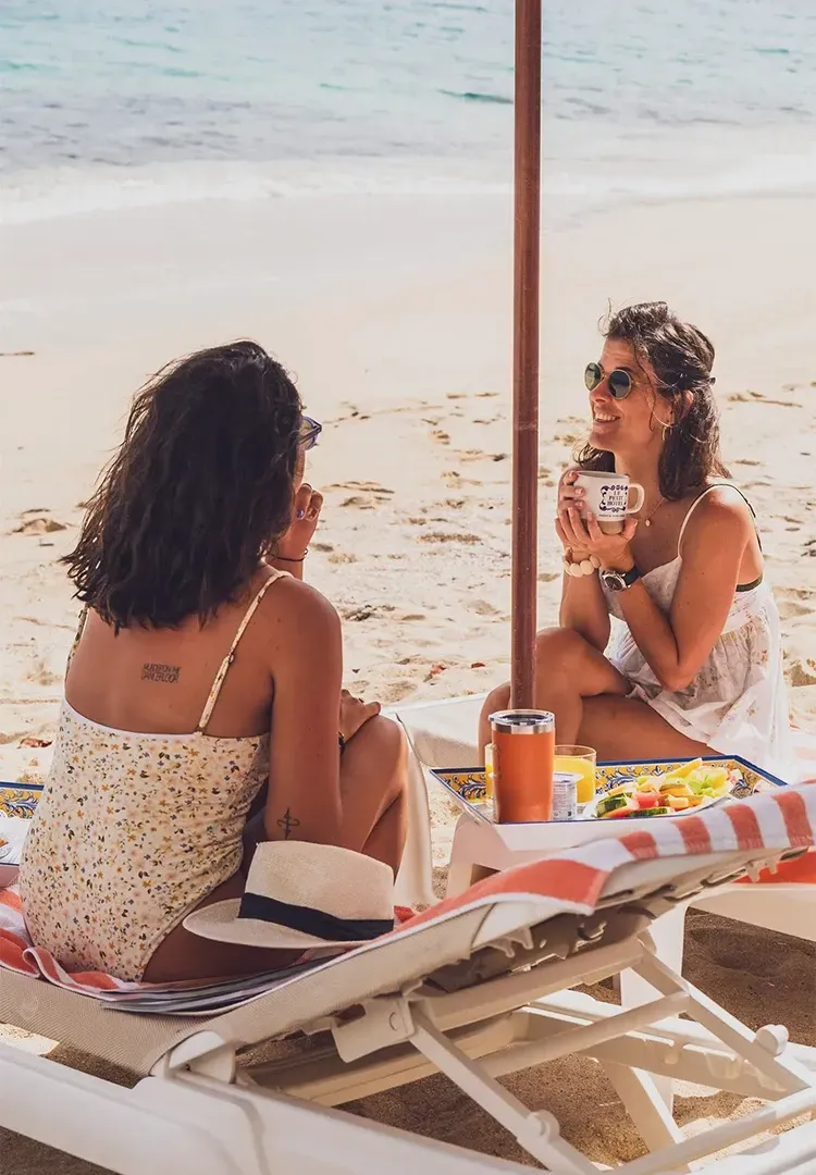 Two women on beach lounge chairs, enjoying drinks and food under an umbrella. Bright sunlight, turquoise water.