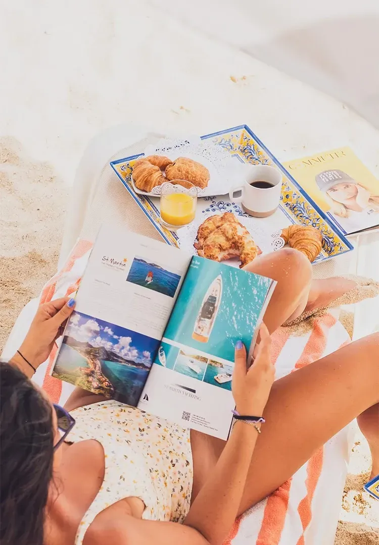 Woman on beach reads magazine, tray with pastries, coffee.