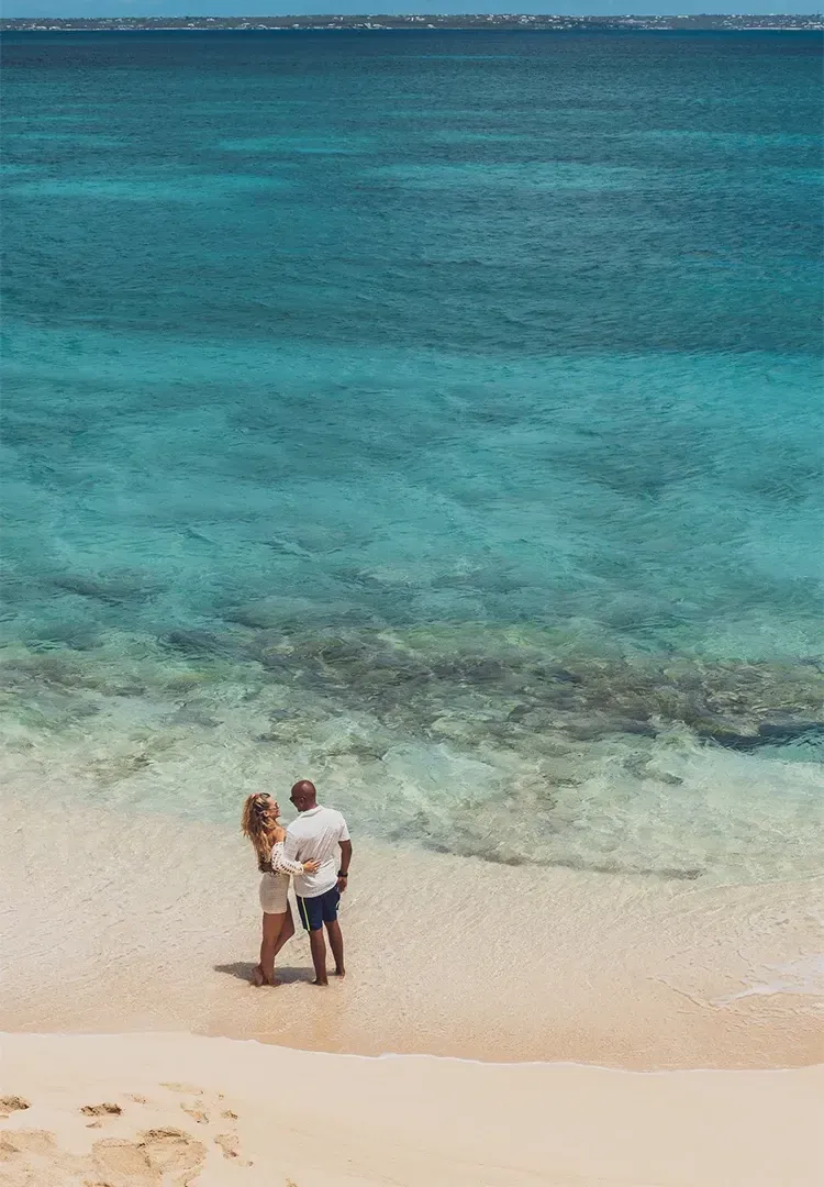 Couple standing in shallow turquoise water at a beach, facing the ocean.