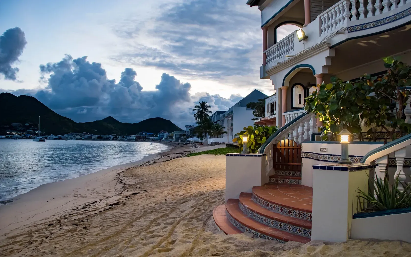 Beachfront villa at dusk, with a sandy shore, calm water, and mountains in the background.