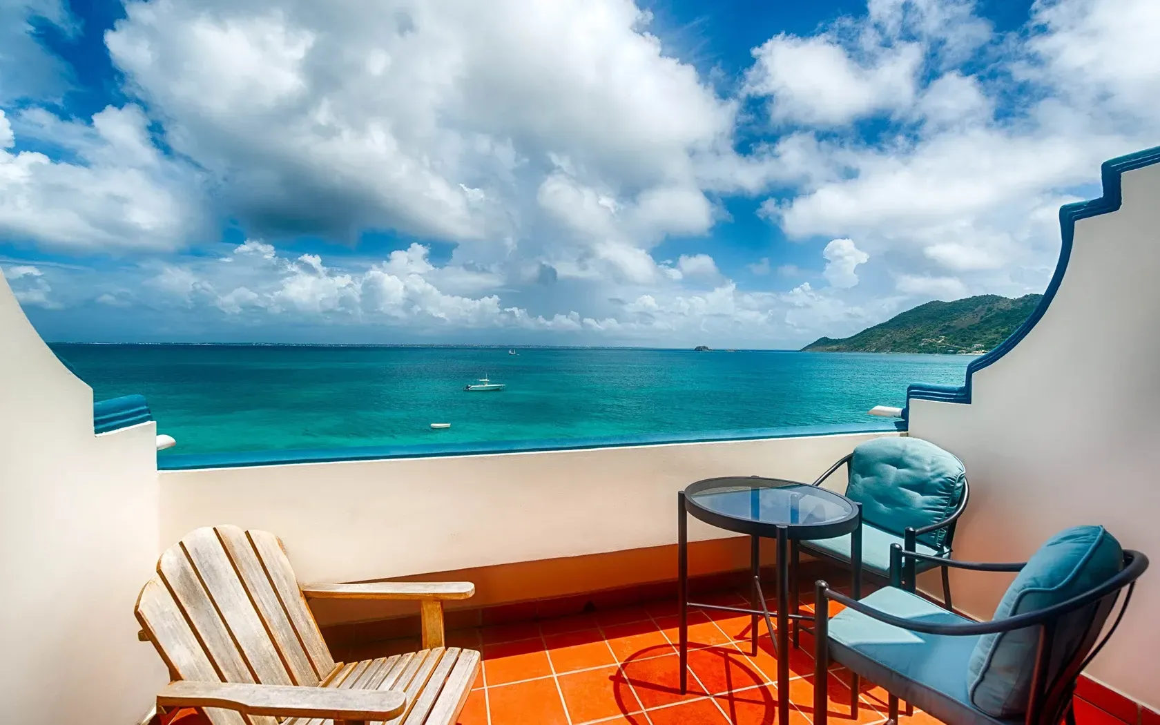 Balcony with ocean view: wooden chair, table, blue chairs, turquoise water, cloudy sky.