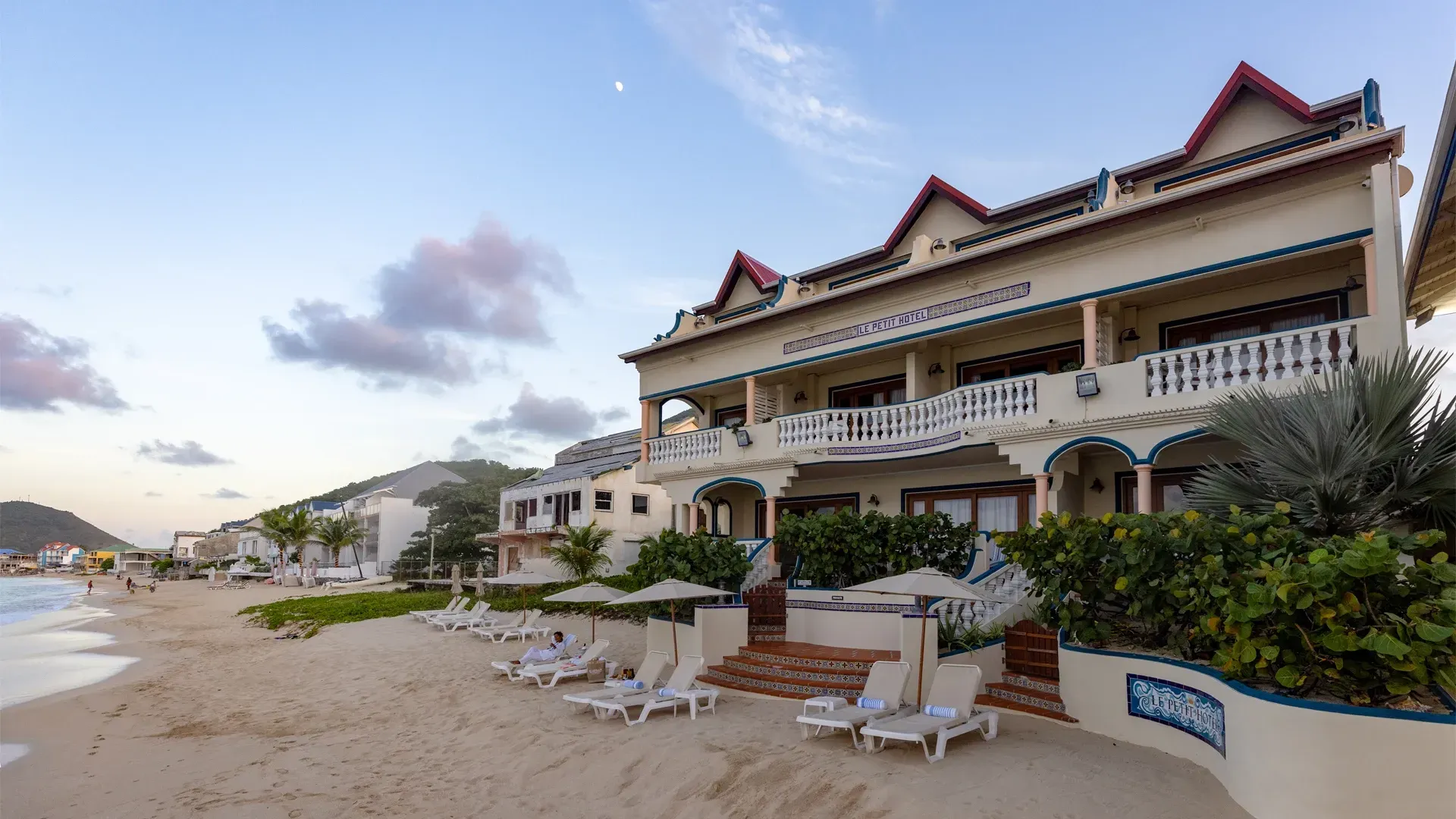 Beachfront hotel with lounge chairs, tan sand, and blue sky.