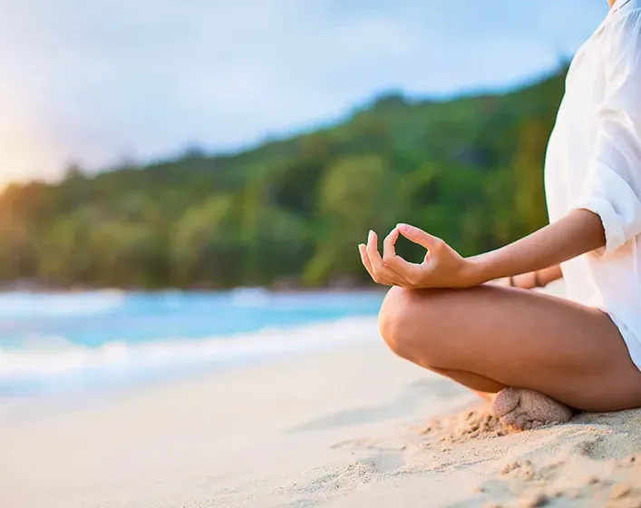 Person meditating on a sandy beach with ocean and green hills in the background.