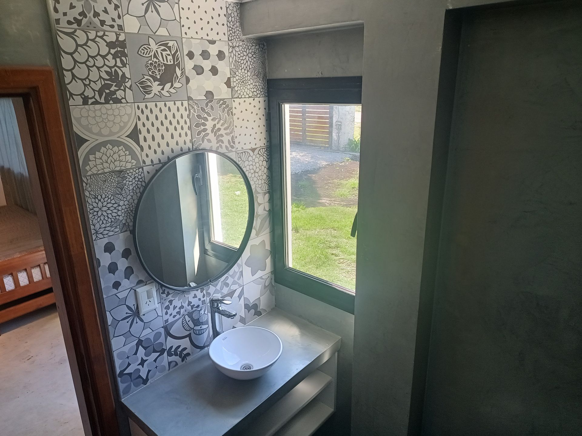 Baño con papel pintado estampado, espejo redondo, lavabo blanco y pequeña ventana con vista al césped.