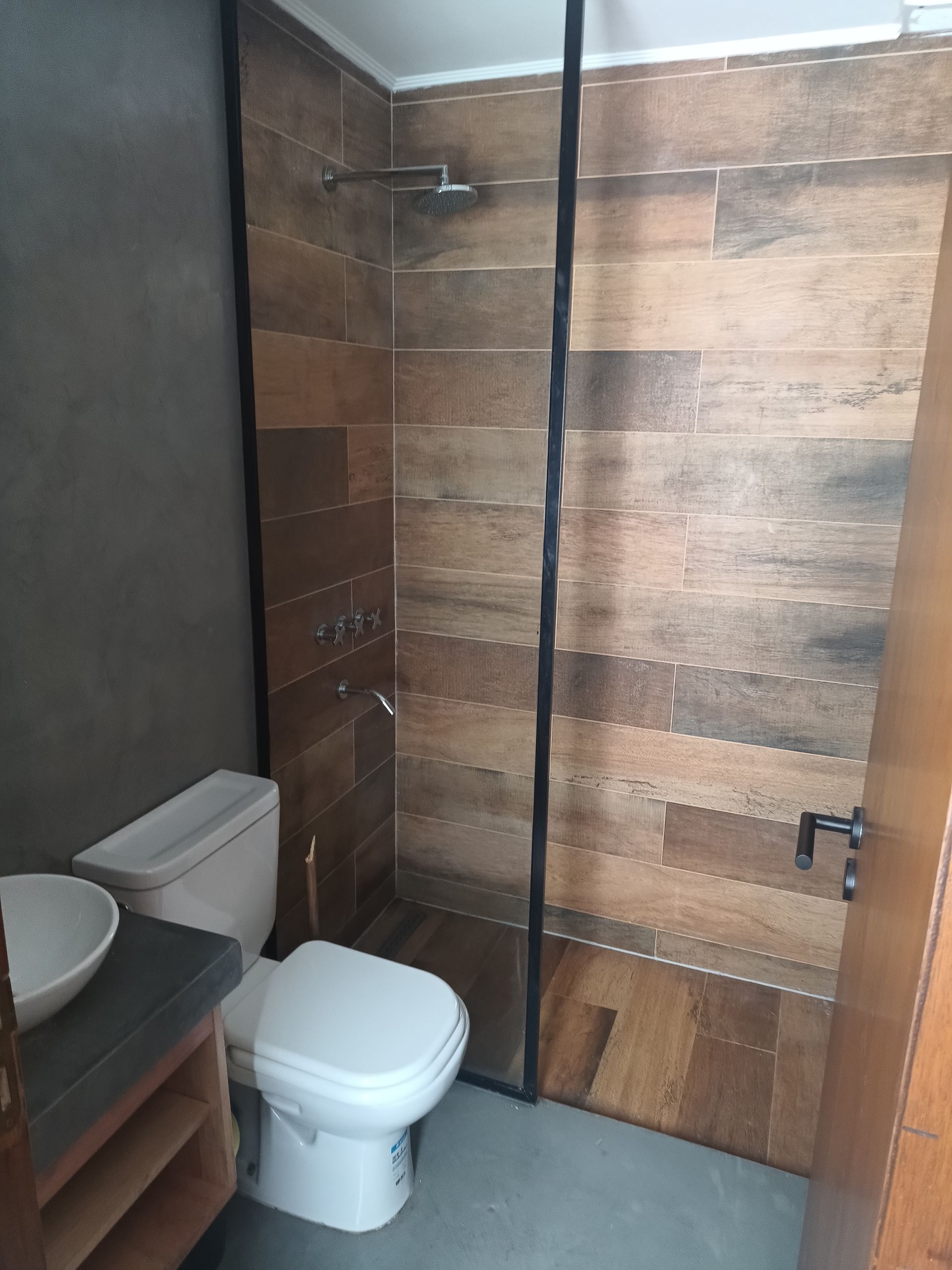 Baño con azulejos imitación madera, mampara de ducha de cristal y WC.