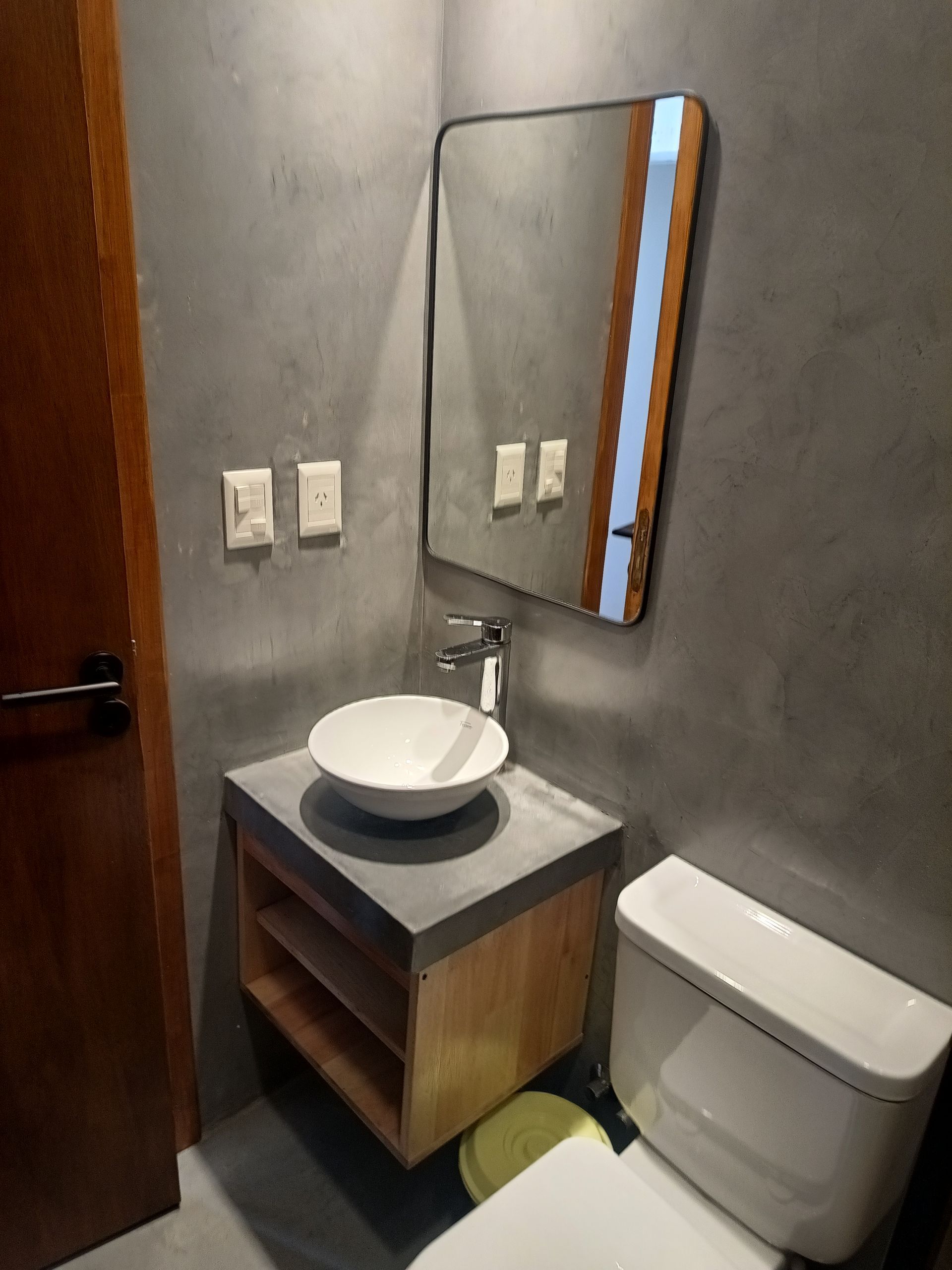 Baño con tocador flotante de madera, lavabo blanco, espejo rectangular y sanitario.
