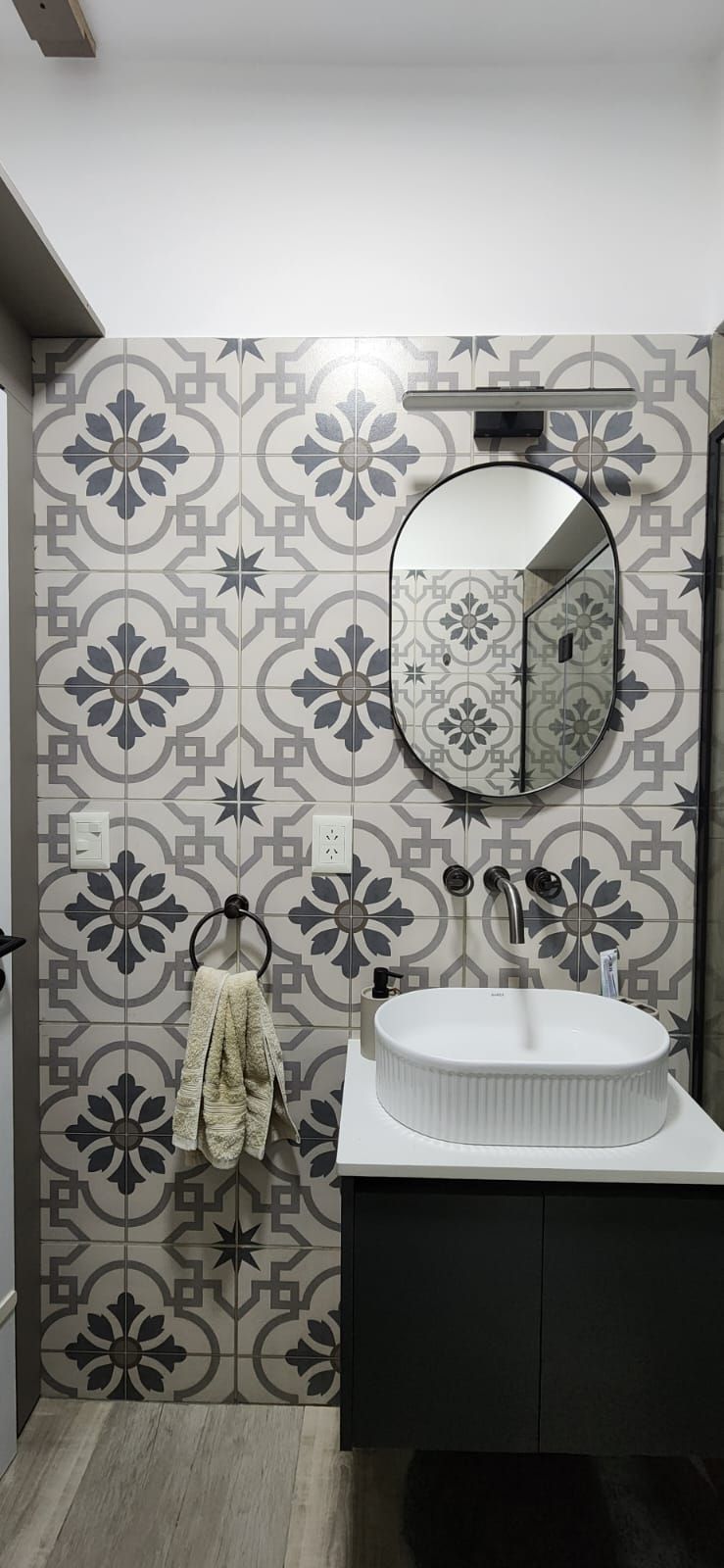 Baño con azulejos estampados en gris y blanco, espejo ovalado, mueble oscuro y lavabo blanco.