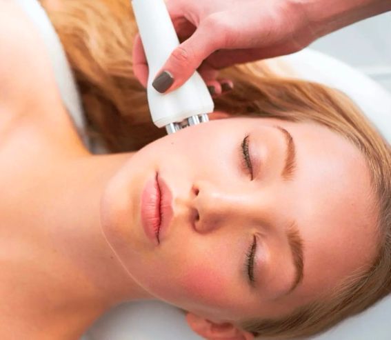Woman receiving facial treatment with a handheld device on cheek; eyes closed, fair skin.