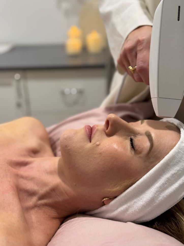 Woman receiving facial treatment; white headband, medical device pointed at face. Beige setting.