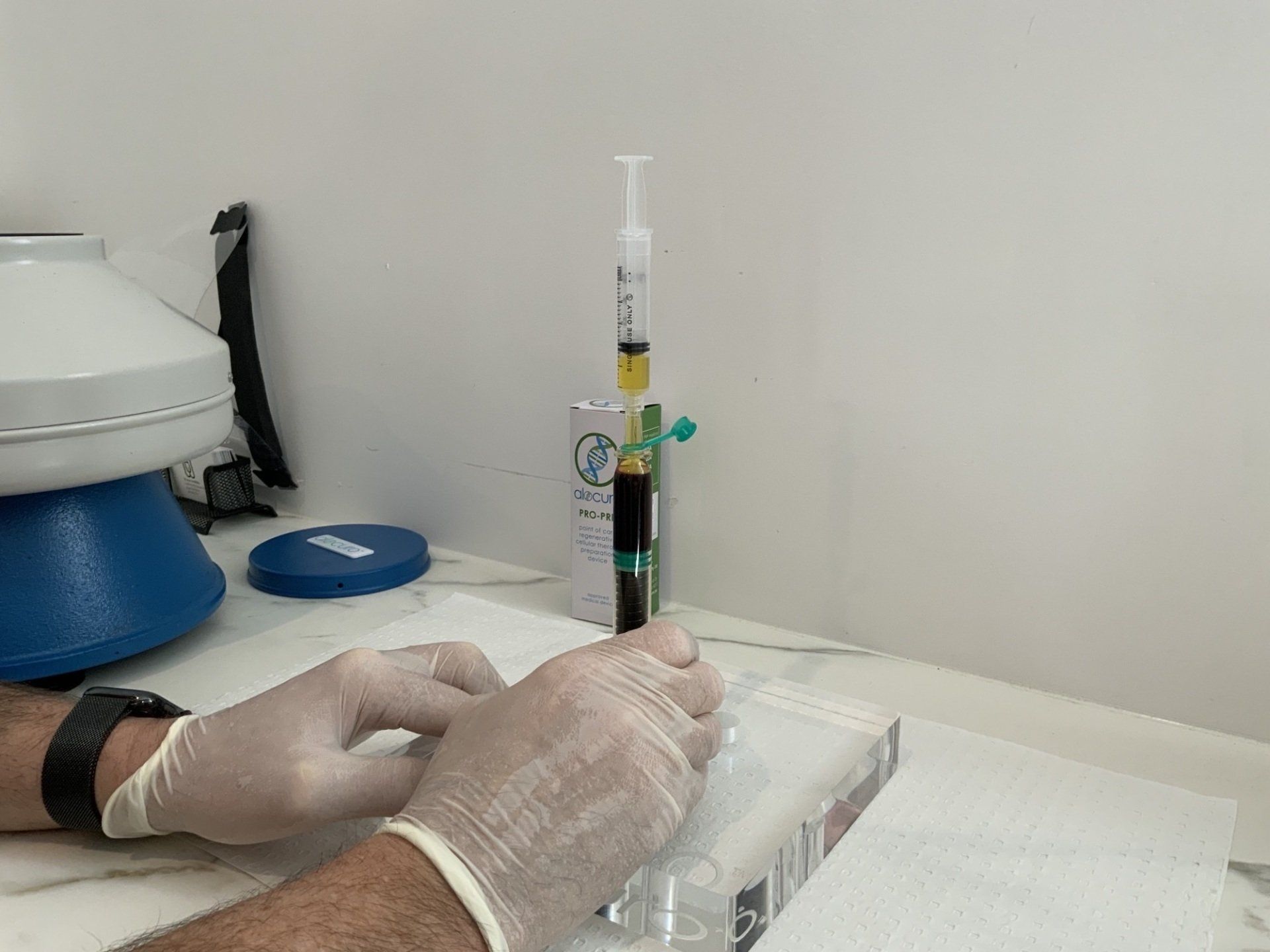 A person wearing gloves is holding a syringe in front of a centrifuge.