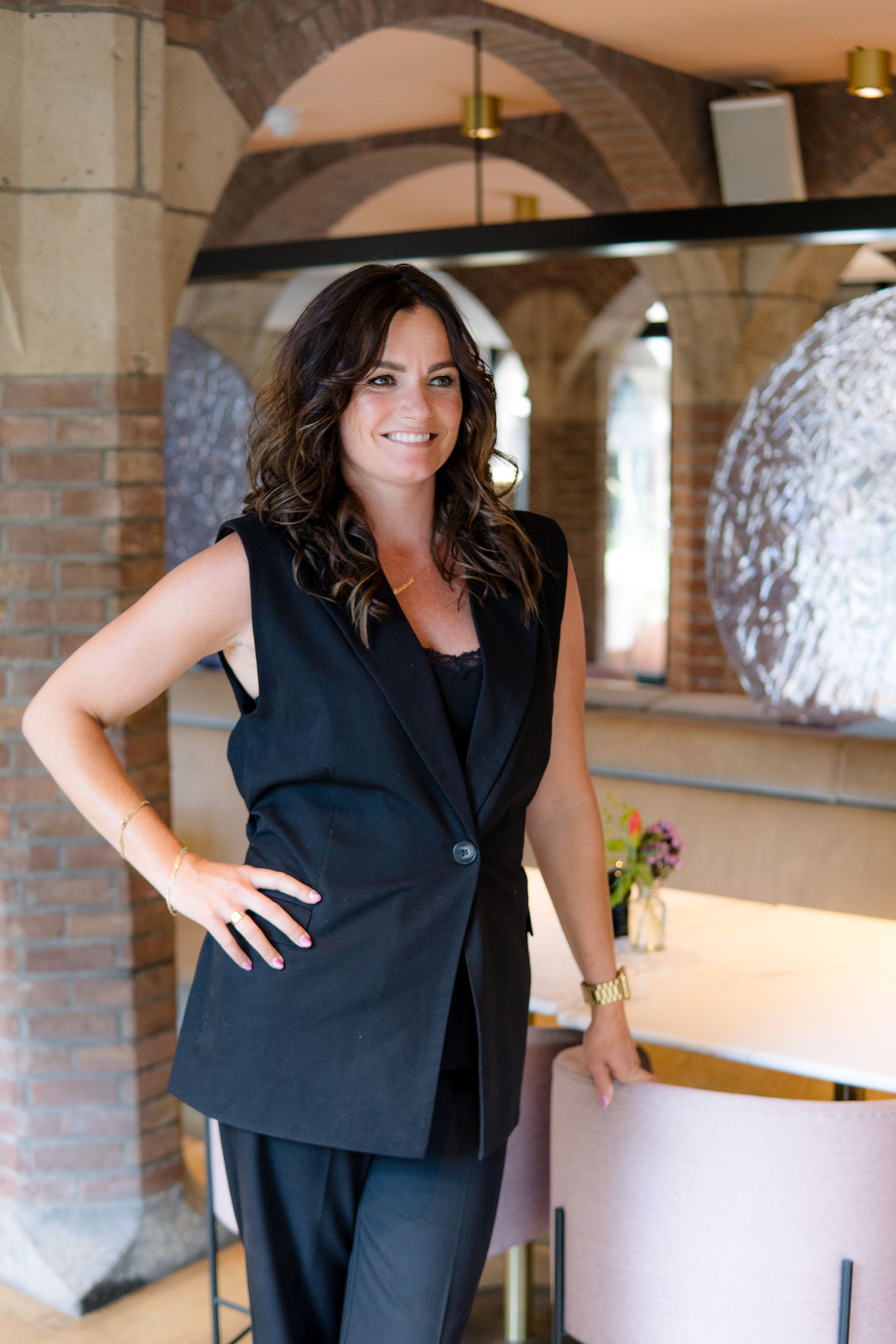 Een persoon in een wit shirt en spijkerbroek zit op stenen trappen die naar een bakstenen gebouw leiden en lacht naar de camera.