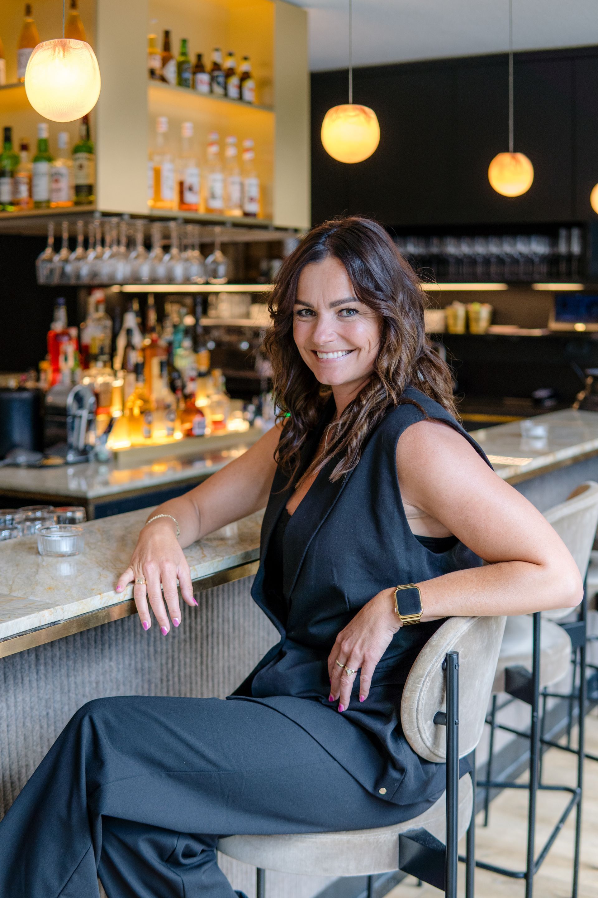 Een glimlachende persoon in een donker vest en een donkere broek zit aan de bar in een goed verlicht, modern restaurant.