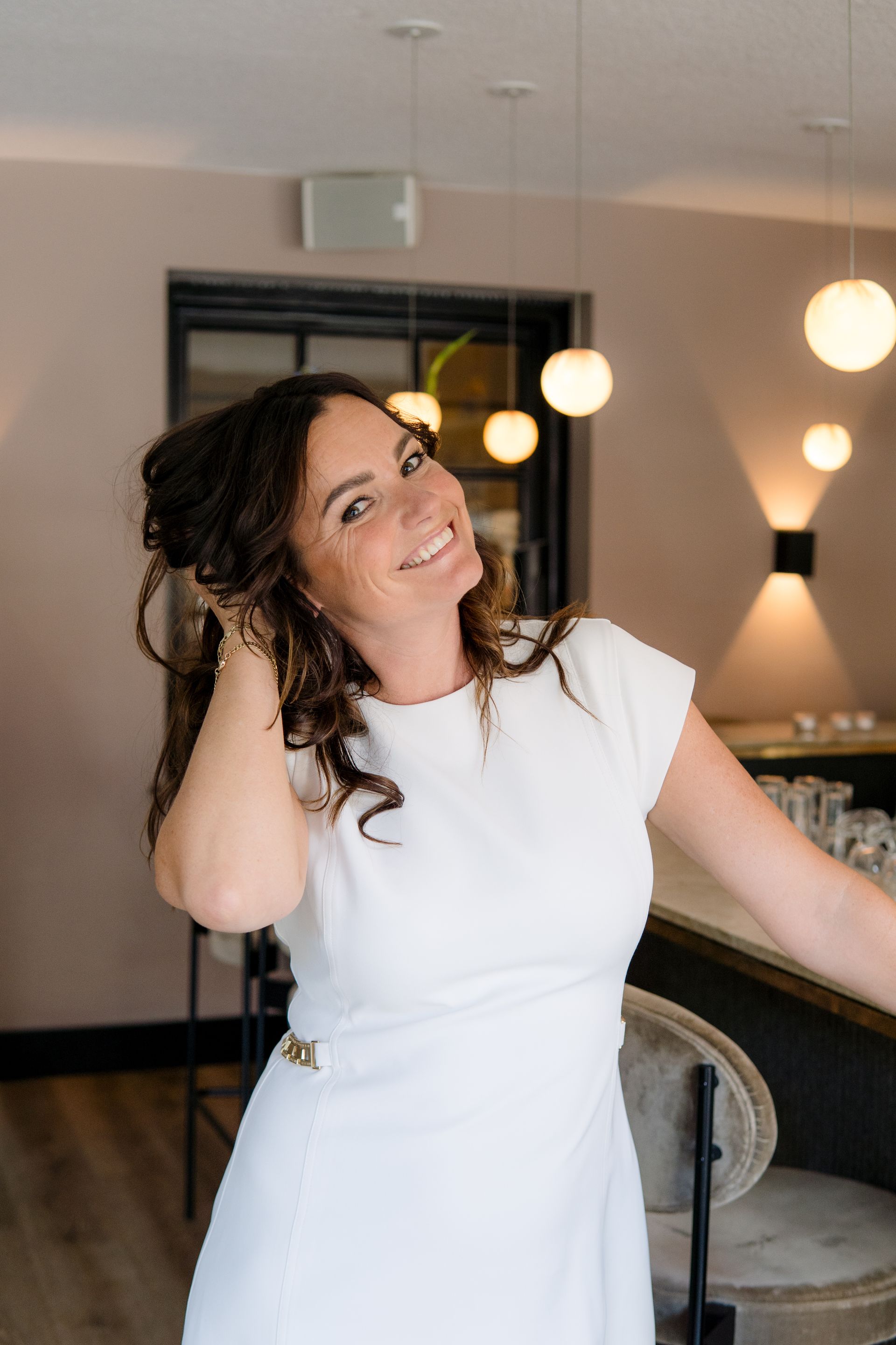 Een vrouw met golvend haar, gekleed in een witte jurk, glimlacht terwijl ze haar hand achter haar hoofd laat rusten in een moderne, verlichte ruimte.
