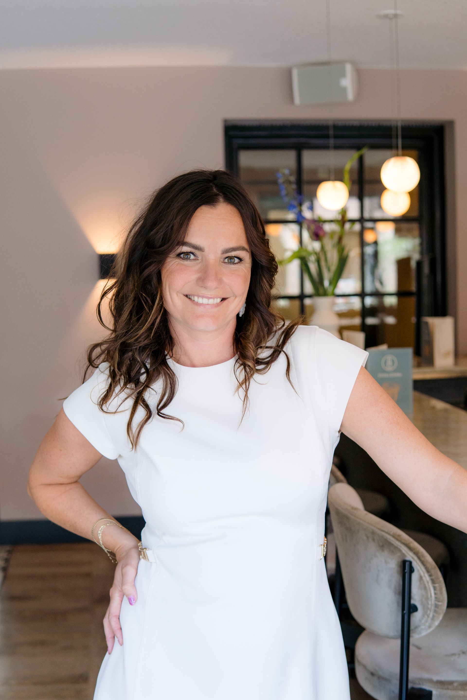 Een lachende vrouw met golvend bruin haar, gekleed in een witte jurk, staat in een restaurant met barzitjes.