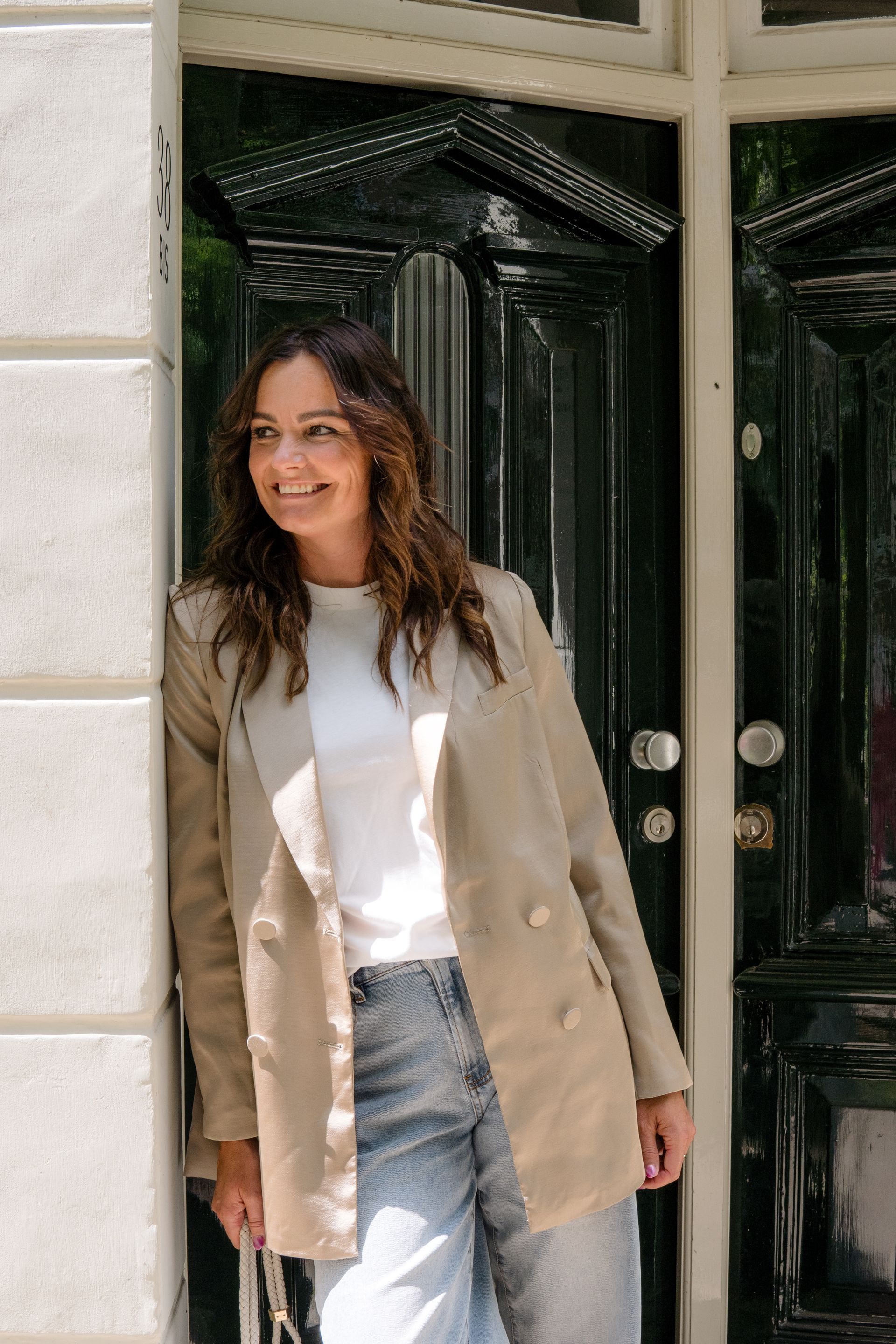 Een persoon glimlacht terwijl hij tegen een witte pilaar leunt naast een donkere deuropening, gekleed in een beige blazer en een blauwe spijkerbroek.