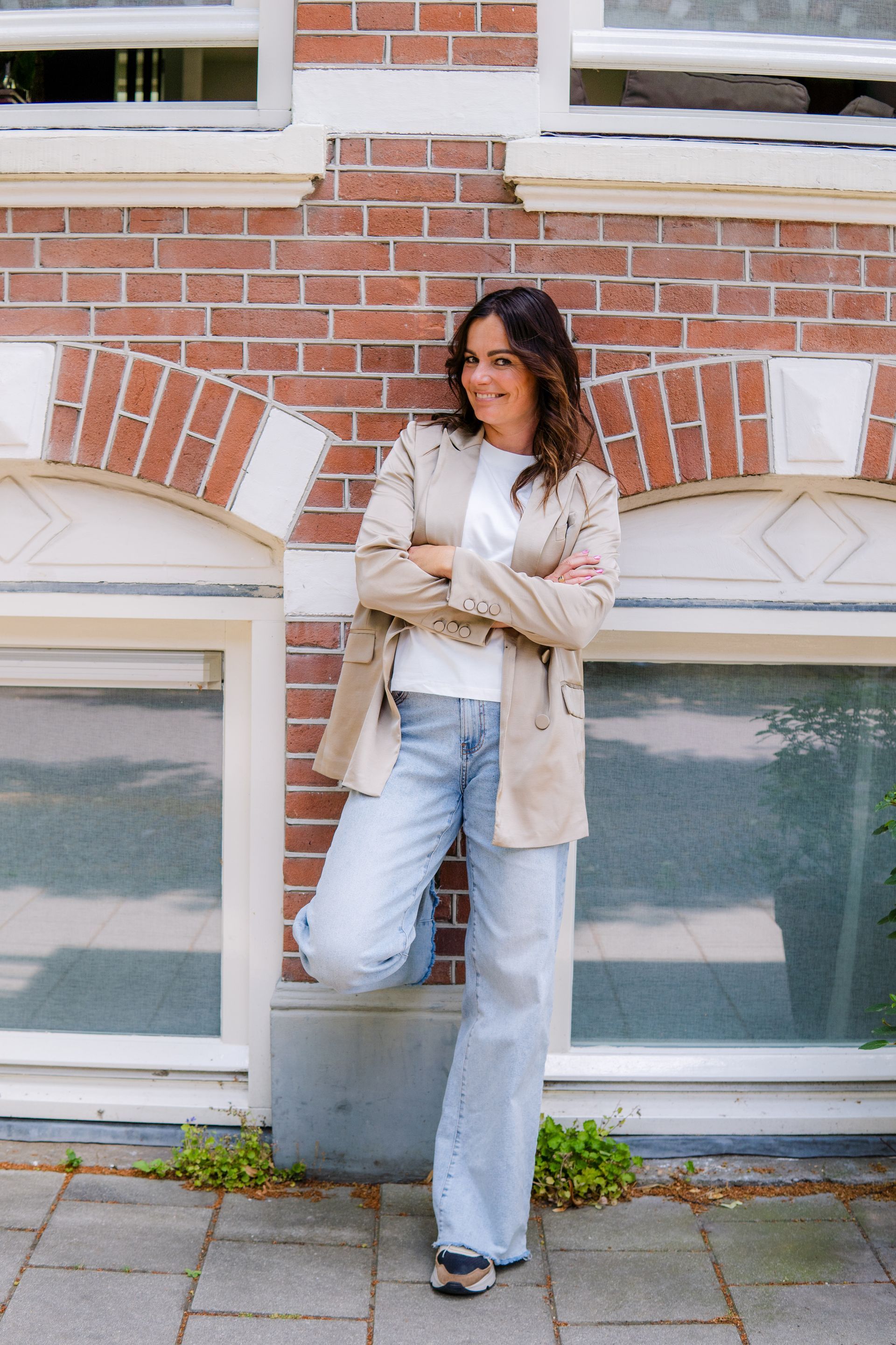 Een persoon met golvend haar, gekleed in een beige blazer, een wit T-shirt en een lichte spijkerbroek, leunt tegen een bakstenen muur bij een raam.