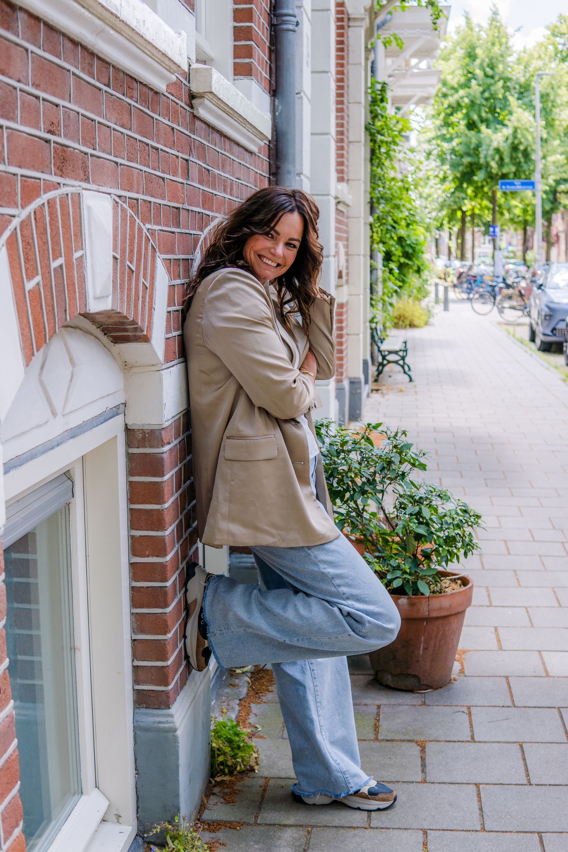 Een glimlachende persoon in een beige blazer en wijde jeans leunt tegen een bakstenen gebouw op een stadsstoep naast een potplant.