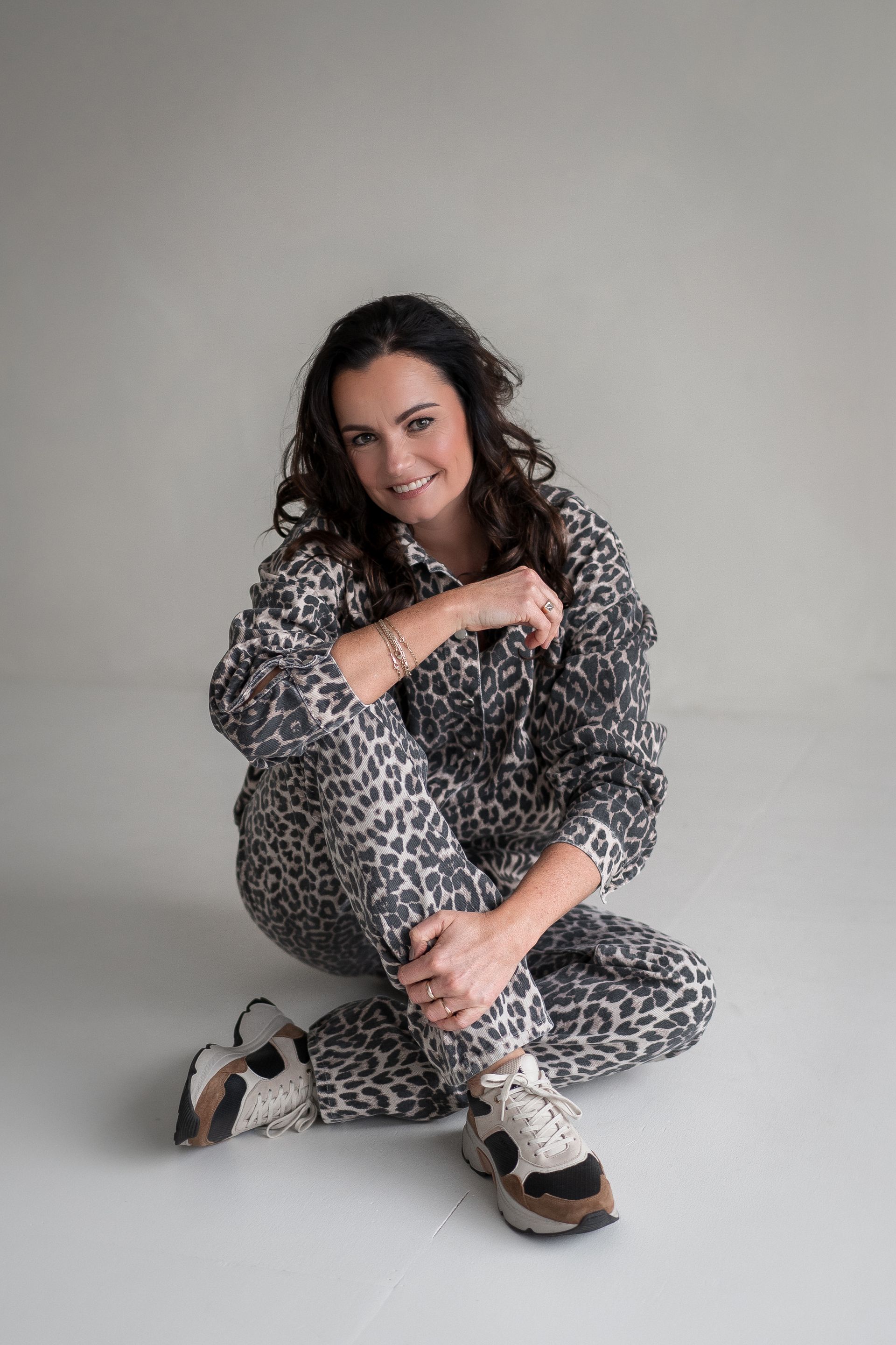 Een persoon met donker haar, gekleed in een grijze outfit met luipaardprint en sneakers, zit op een effen, lichtgrijze vloer.