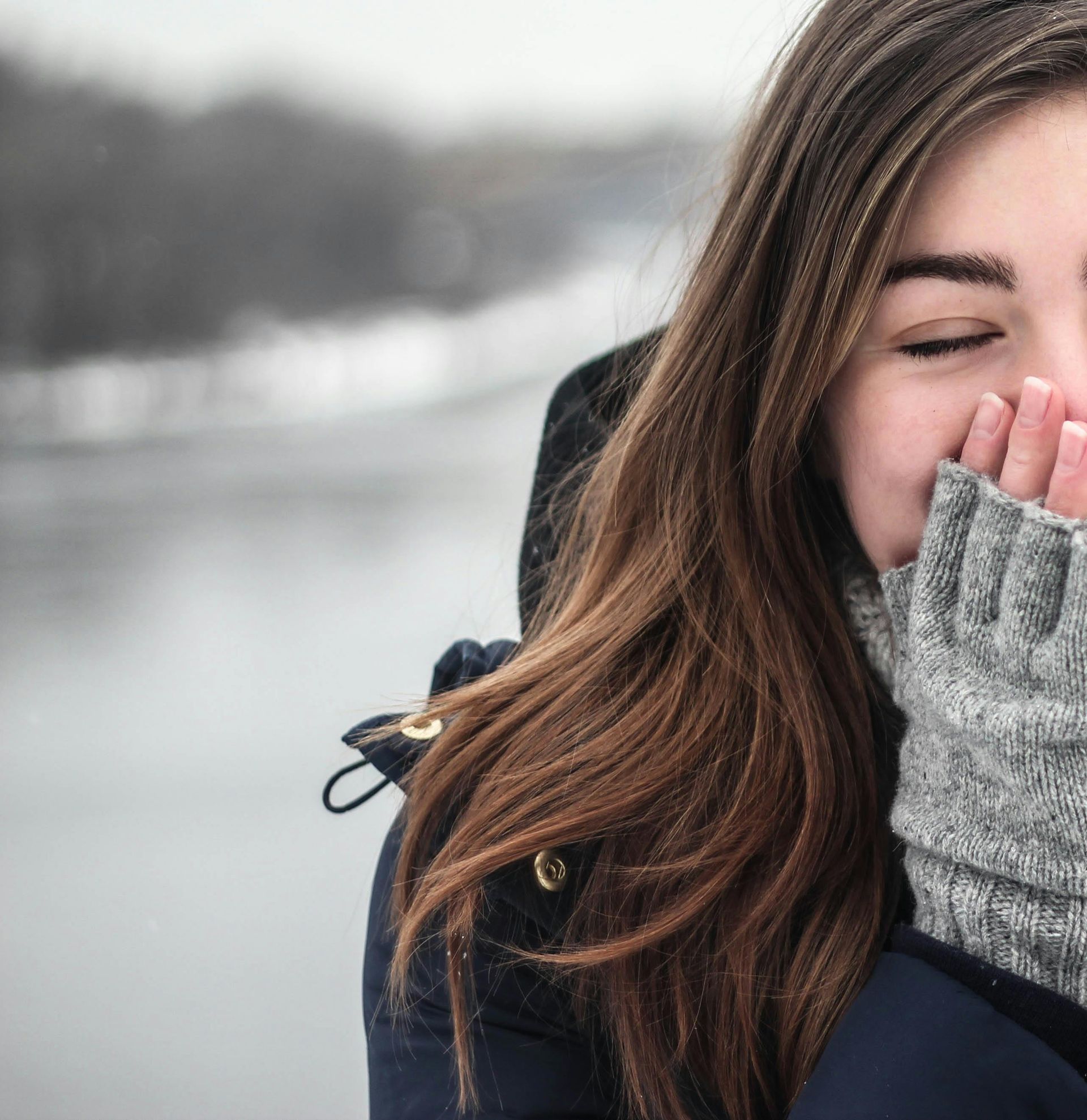 Woman outside, smiling, holding hands up to face, wearing a gray sweater and dark coat, snowy background.