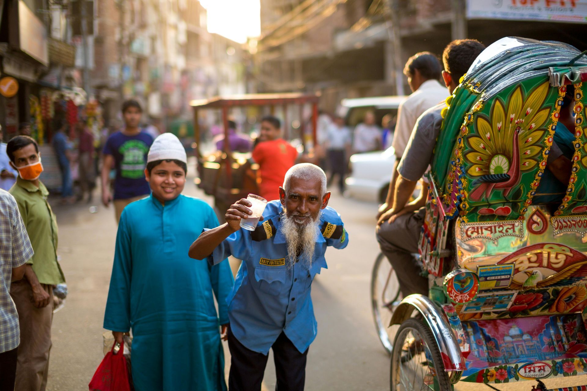 Street scene in Bangladesh: people, a rickshaw, and buildings. An older man smiles, holding a drink, with a boy in the foreground.