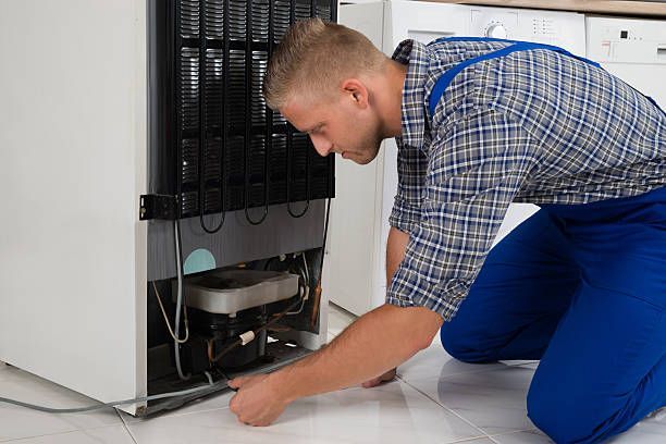 Un hombre está arrodillado en el suelo arreglando un refrigerador.