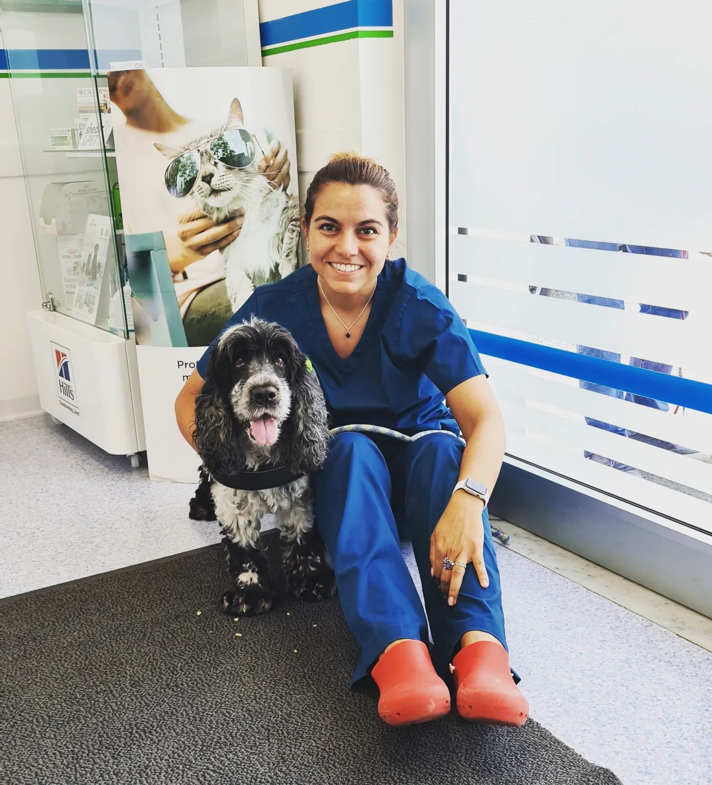 Donna in camice con cane, entrambi sorridenti. Seduti vicino a una finestra in una clinica veterinaria.