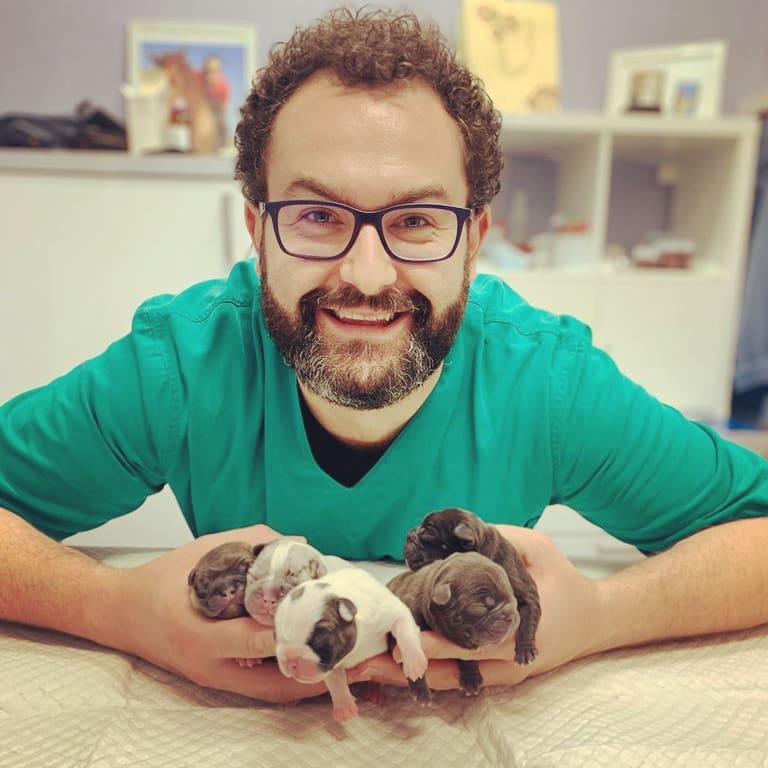 Uomo con gli occhiali, sorridente, tiene in mano una manciata di cuccioli appena nati. Camicia verde, ambientazione interna.