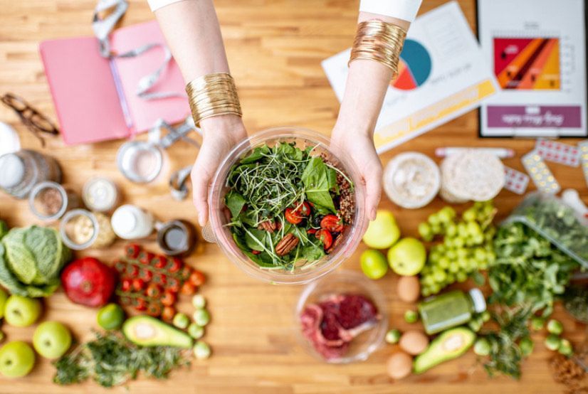 A woman is holding a bowl of salad in front of a table filled with fruits and vegetables.