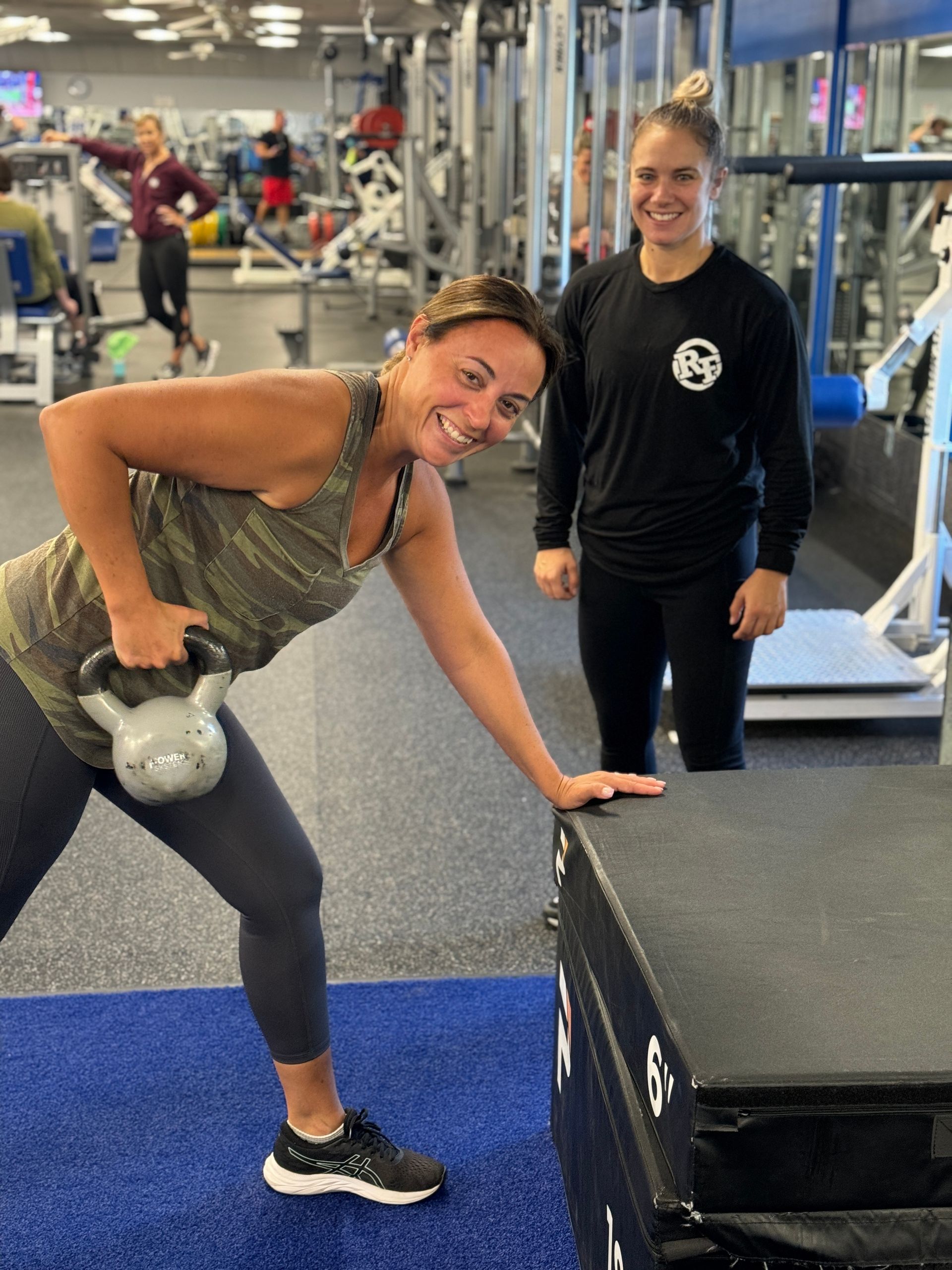 A woman is lifting a kettlebell in a gym while another woman watches.