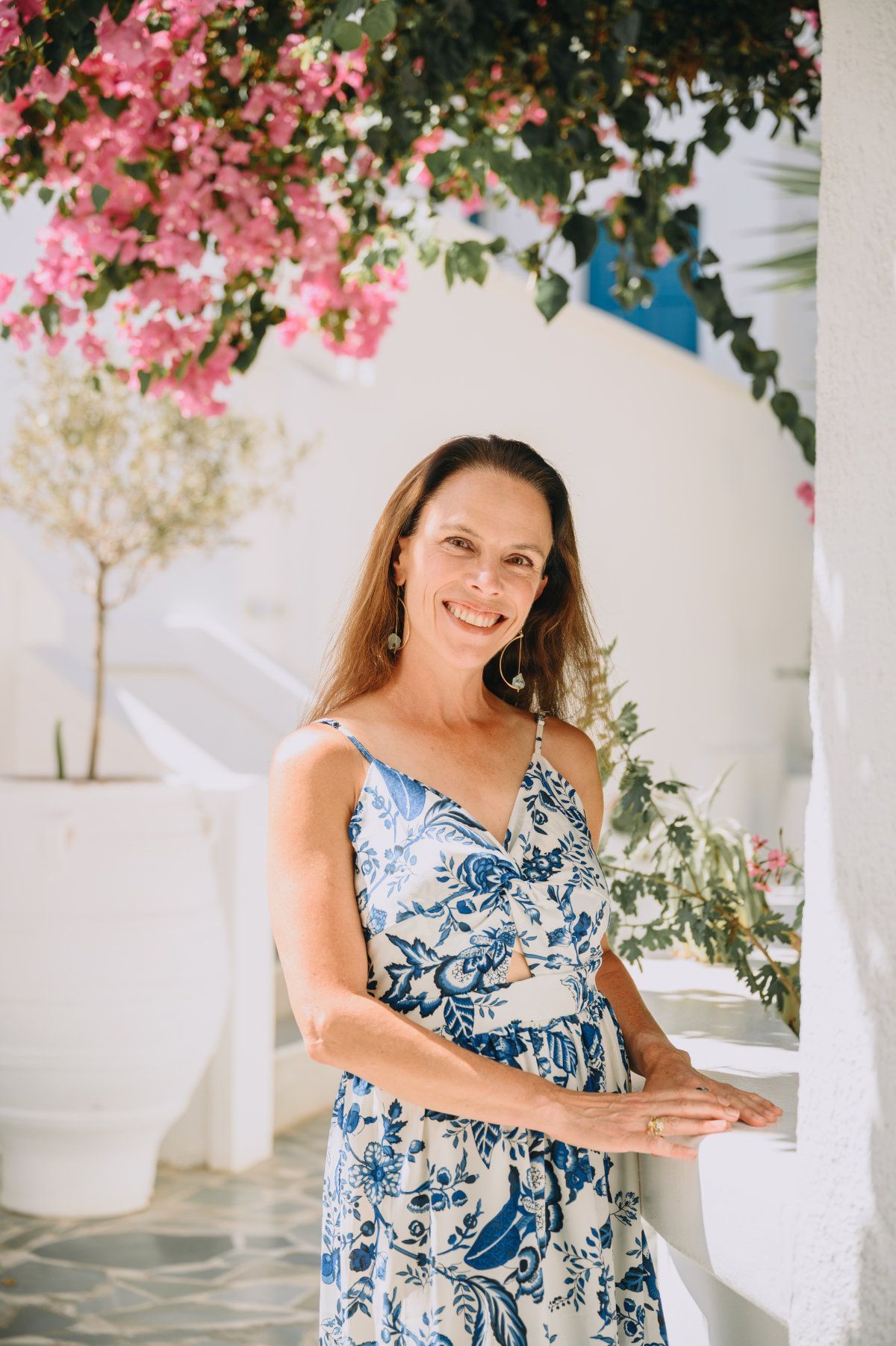 Woman in blue and white dress smiles outdoors, leaning on a white wall. Pink flowers and plants overhead.