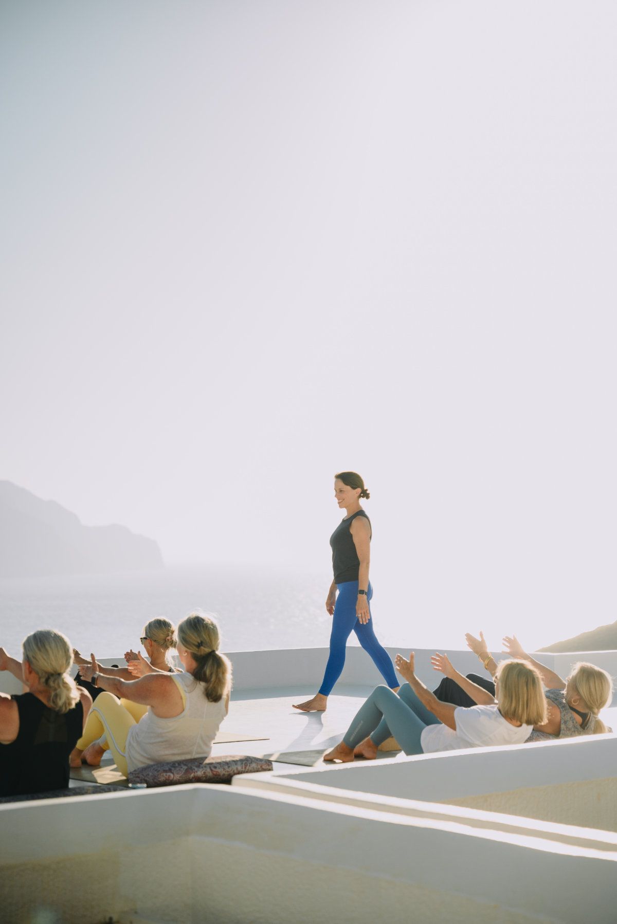 Yoga class outdoors, instructor leading poses. Participants on a white patio with a sunny, mountain backdrop.