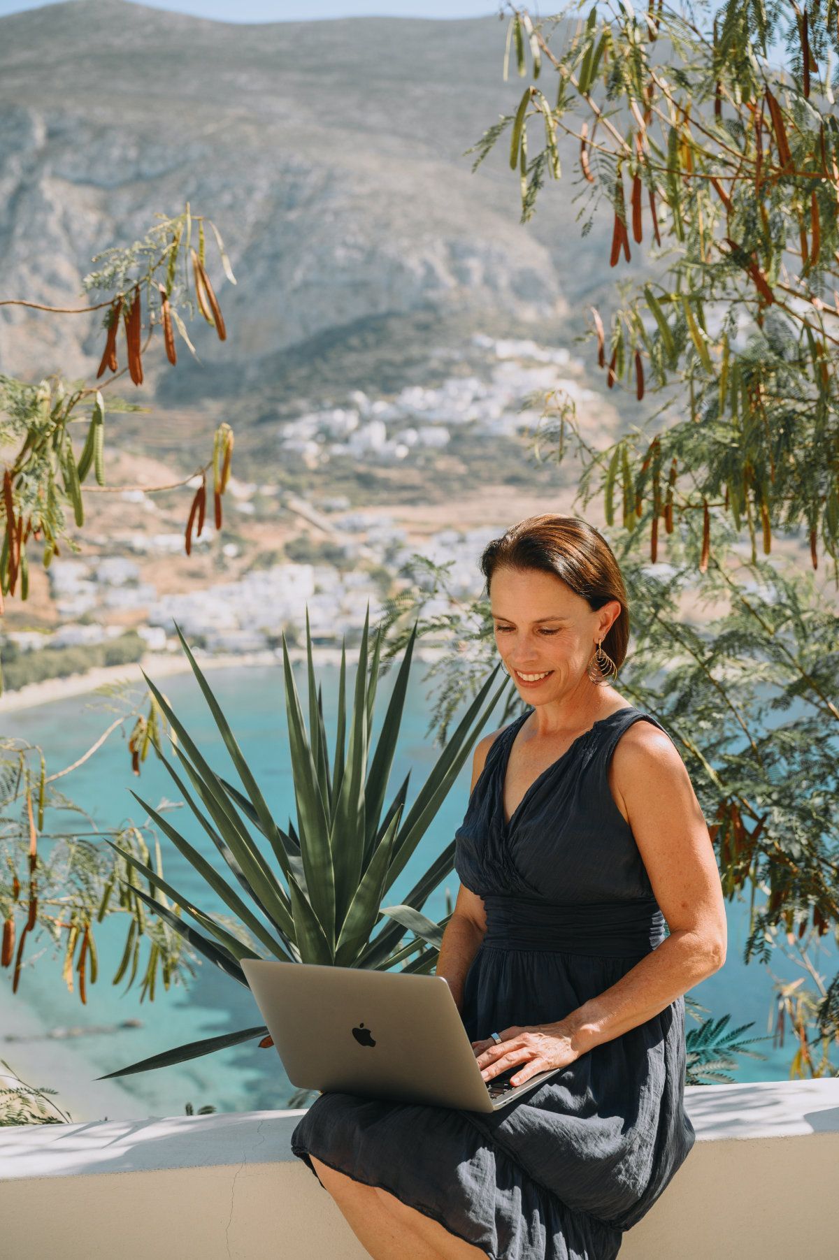 Woman sitting with laptop, smiling, overlooking a coastal village and turquoise water.